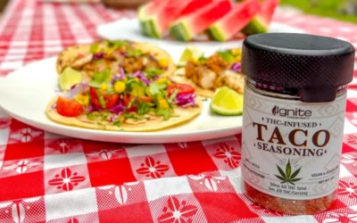 Taco-infused seasoning jar from Ignite Dispensary beside vibrant tacos topped with fresh vegetables, set on a red checkered tablecloth, highlighting innovative THC-infused culinary products.